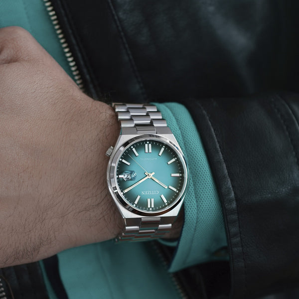 CITIZEN WATCHES Mod. NJ0151-88X displayed on wrist with turquoise dial and silver bracelet, styled with teal shirt and black jacket.