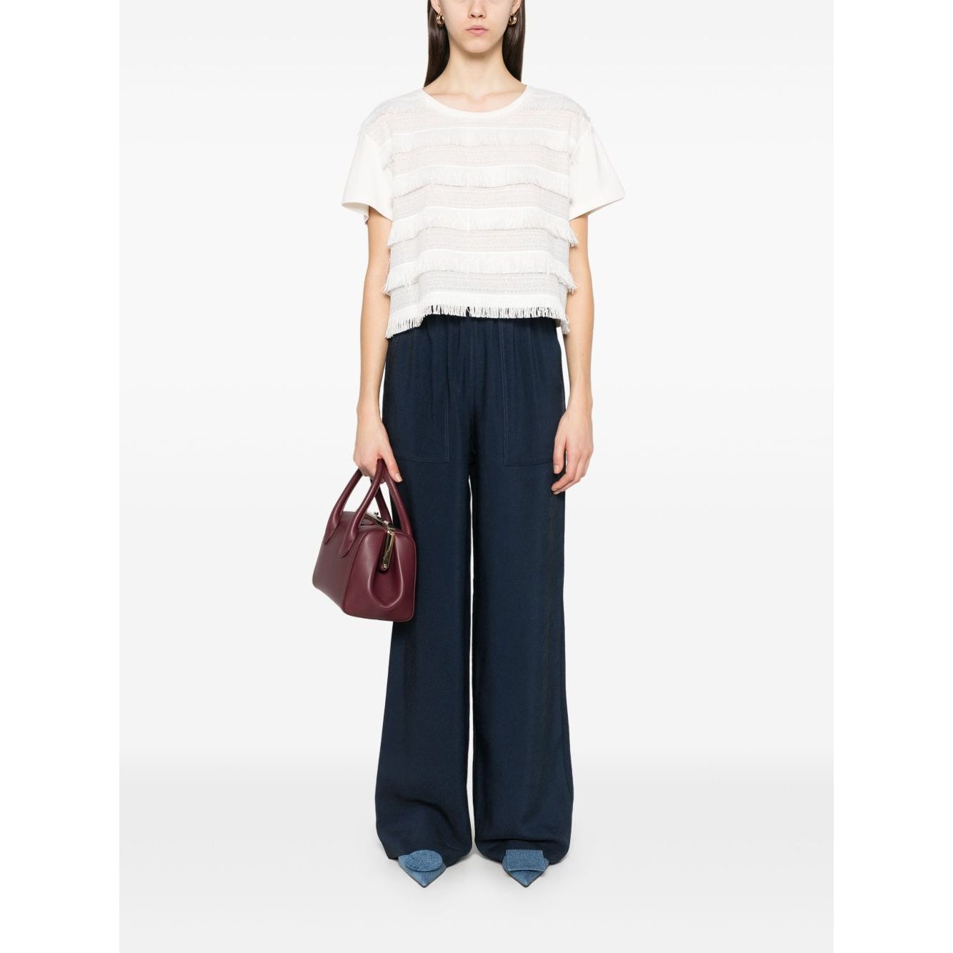 Woman wearing Emporio Armani T-shirt with fringes, white with embroidered logo, paired with blue wide-leg pants, holding a burgundy bag.